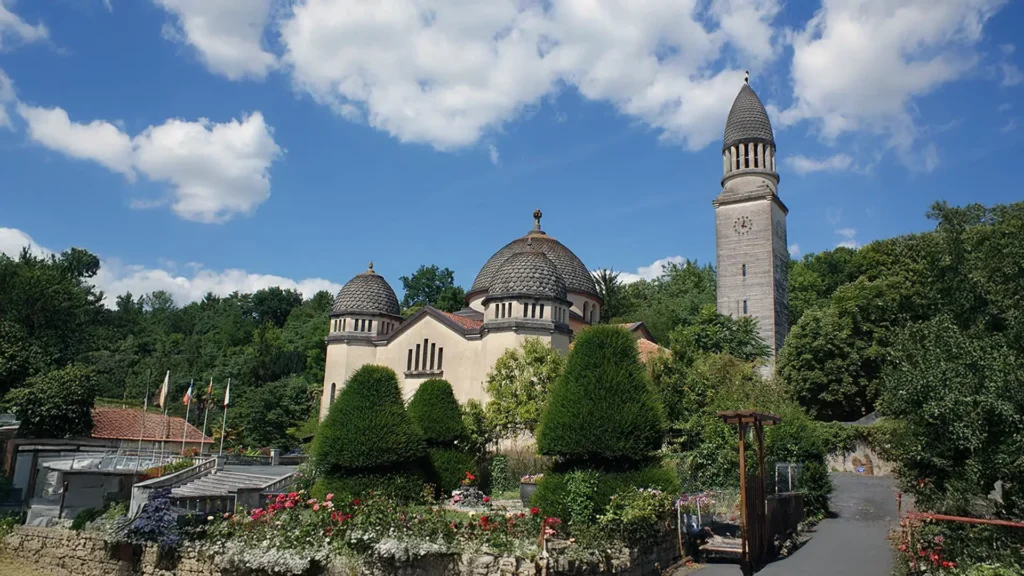 A wide shot of a cream-colored, Neo-Byzantine style church under a bright blue sky with scattered white clouds. The church features two dark, tiled domes and a tall, slender bell tower topped with a conical dome on the right. In the foreground, a manicured garden is filled with low stone walls, vibrant red and white flowers, and shaped conical evergreens. A narrow asphalt pathway runs along the right side next to a wooden pergola structure. The building is surrounded by dense green forest on a sloped hillside in the background.