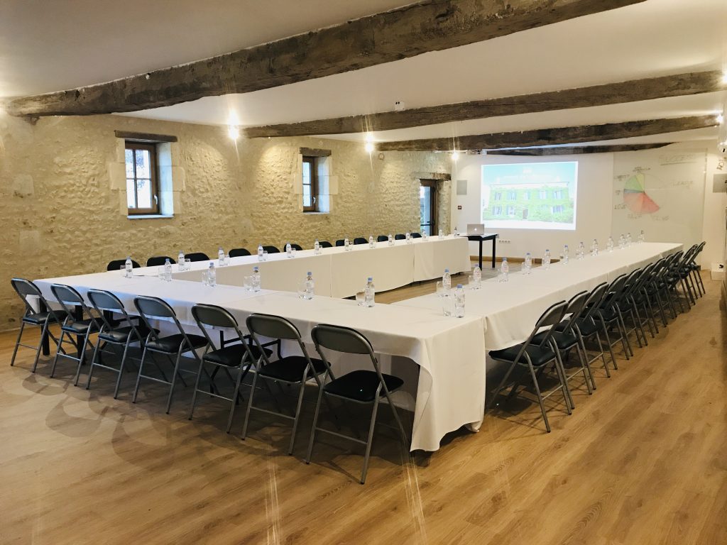 A wide, eye-level shot captures a large conference room set up for a meeting inside a rustic building with exposed stone walls and thick wooden ceiling beams. Long tables draped in white tablecloths are arranged in a large U-shape, lined with numerous black folding chairs along the outside. Each seating position is set with a clear glass and a plastic water bottle. At the far end of the room, a projector screen displays an image of a green, ivy-covered house next to a whiteboard featuring a colorful pie chart. Two small, deep-set windows on the left wall allow a glimpse of daylight into the brightly lit space.